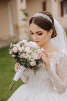 brunette bride in a lacy white dress with a long train, posing with a bouquet of white and pink flowers, against the background of trees. The veil is in the air. Beautiful hair and makeup. photo