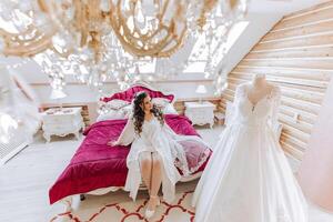 A happy bride is preparing for her luxurious wedding in a hotel room, with a wedding dress on a mannequin nearby. Portrait of a woman with fashionable hair, makeup and a smile in a dressing gown. photo