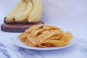 Crispy banana chips on a white plate with fresh bananas. photo