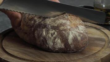 Cutting slice of homemade Crusty rye bread with a sharp knife on wooden board, closeup slow motion video