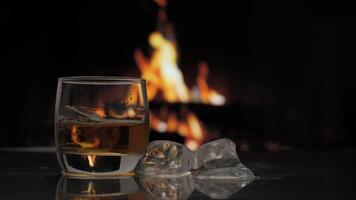 Glass of whiskey, scotch or bourbon with ice stands on a table against background of a fireplace with a flame. Alcoholic drink and home comfort concept. video