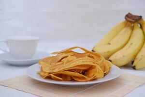 Crispy banana chips on a white plate with fresh bananas. photo