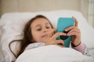 Focus on smartphone in hands of blurred little girl, addicted to digital gadget and online video games, lying on the bed photo