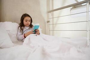Little kid girl, addicted to digital gadgets, playing online video games while lying on the bed covered with white duvet photo