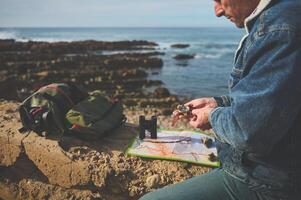 relajado hombre caminante turista viajero mirando para direcciones con un Brújula, sentado en el rock en el atlántico playa. foto