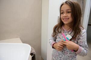 Little child girl in pajamas, holding kids toothpaste and her pink toothbrush, standing in the bathroom at home photo