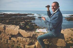 sonriente turista viajero sentado en un rock por mar cerca su mochila, participación prismáticos, sonriente mirando a el cámara foto