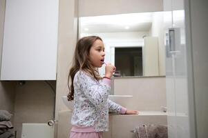 pequeño niño niña cepillado su dientes, en pie en el gris minimalista hogar baño. dental cuidado. oral higiene. Copiar espacio foto