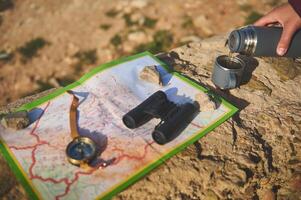 Hand holds thermos flask and pours hot tea drink into stainless steel mag, near a map, compass and binoculars on a cliff photo