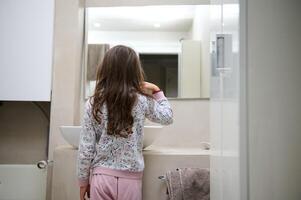 Rear view child girl standing at washbasin in the home bathroom and brushing her teeth, looking at herself in the mirror photo