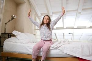 caucásico linda pequeño niña despertar arriba en el mañana, extensión su brazos hacia arriba, sonriente a cámara, sentado en el cama foto