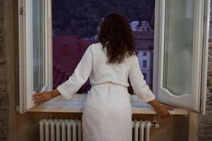 Rear view of a young woman in white bathrobe, opening windows in the living room, enjoying dawn in the early morning photo