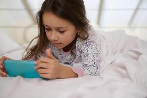 Caucasian lovey little child girl in nightwear, watching cartoons on mobile phone before sleeping, lying down on her bed photo