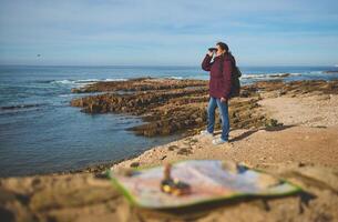 Woman with a backpack, looking in binoculars standing on top of a cliff, enjoying a beautiful view of the Atlantic ocean photo