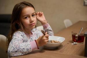 auténtico retrato de un hermosa caucásico pequeño niño niña disfruta comiendo cereal para Mañana desayuno con apetito. foto