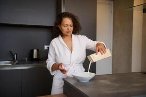 desayuno concepto. contento soñoliento mujer en un túnica, torrencial Fresco Leche desde Bosquejo tetra paquete botella dentro cuenco con cereales foto