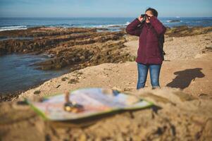 Young adult traveler woman adventurer looking into the distance through binoculars, standing on the top of cliff by sea photo