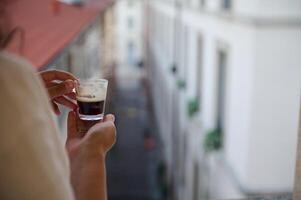 Closeup of woman's hands hold cup of freshly brewed espresso, standing by windows and admiring the beautiful city view photo