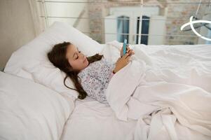 adorable elemental años niño, pequeño niño niña utilizando teléfono inteligente, acecho películas mientras acostado en el cama en el Mañana. foto