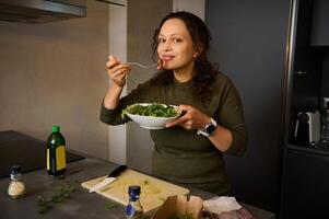 Pretty young woman standing at kitchen table, holding a fork with salad near her face, smiles cutely looking at camera photo