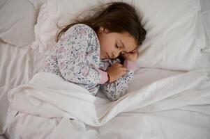 parte superior ver hermosa encantador pequeño niño niña en elegante pijama, dormido en su cama, cubierto con blanco limpiar sabanas foto