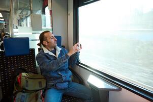 Cheerful young adult Caucasian man in casual denim, gazing out the window during his morning commute on an urban light rail train, smiling and taking photo of nature out the window on his smartphone