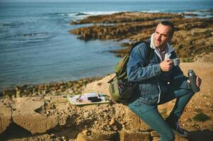 Happy adventurer sitting by Atlantic beach, smiles looking aside, holds a thermos, enjoying his coffee break outdoor photo