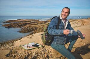 Happy male tourist sitting on a rock by sea, holding a thermos bottle and pouring some coffee into a steel mug photo
