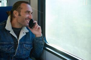 hermoso caucásico joven hombre viajero diario al trabajo hablando en móvil teléfono y mirando fuera el ventana mientras de viaje por tren foto