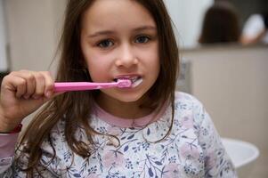 de cerca retrato de un adorable pequeño niño niña cepillado dientes, mirando a el cámara. dental salud y oral higiene foto