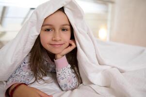 Close-up portrait of a Caucasian beautiful little kid girl lying on the bed under white duvet, smiling looking at camera photo