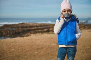 Adorable child girl drinking warm tea thermos cup, looking at camera, standing on the beach, splashing waves background photo