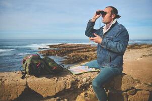 Young man with backpack, looking in binoculars sitting on top of cliff, enjoying a beautiful view of the Atlantic ocean photo