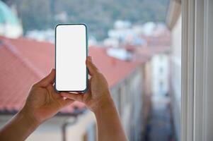 manos sostener teléfono inteligente con blanco blanco Bosquejo pantalla, en contra fondo de hermosa ciudad con casas con rojo techo losas foto