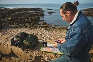 Handsome relaxed young man, hiker tourist sitting on he cliff and orienteering in the nature with compass in the hand photo