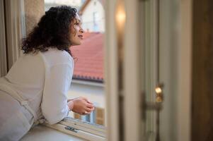 Happy delightful young woman in a white robe looks out the window enjoying the beautiful view of the city of Como photo