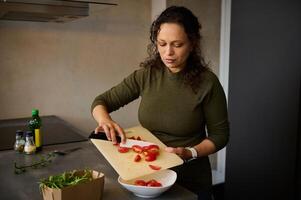 Multiracial woman chef holding kitchen knife, slicing, pouring red tomatoes from cutting board into white bowl for salad photo