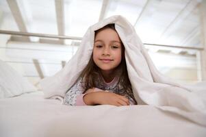 de cerca retrato de un pequeño niño niña 6 6 años antiguo acostado en el cama debajo blanco cobija mientras despertar arriba en el Mañana foto