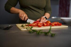 Close-up hands of female chef using kitchen knife, chopping tomatoes on wooden cutting board, standing at kitchen table photo