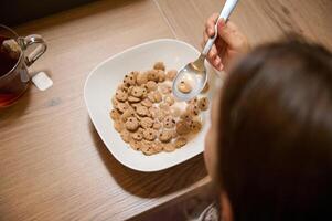 Top view little girl holding spoon full of healthy wholesome oat cereals with milk, taking her breakfast with appetite photo