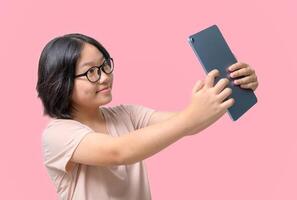 Cute asia girl wearing glasses takes a selfie of herself with a tablet on pink background, photo