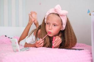 el niña mentiras en el cama y pinturas su labios. el niño mira en el espejo y aplica labio bálsamo. foto