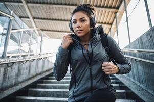 AI generated A women with headphone listen music in a grey hoodie and black backpack running down for concrete stairs. Concept healthy sport lifestyle. photo