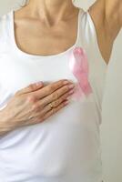 Shot of the woman against the white wall in the white top with pink ribbon, as a symbol of a breast cancer awareness, performing self examination of the breasts, looking for abnormalities. Concept photo