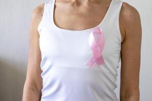 Shot of the woman against the white wall in the white top with pink ribbon, as a symbol of a breast cancer awareness. Concept photo