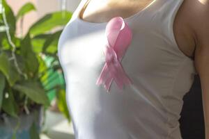 Shot of the woman against the white wall in the white top with pink ribbon, as a symbol of a breast cancer awareness. Concept photo