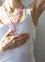 Shot of the woman in the white top, with pink ribbon on her neck as a symbol of breast cancer awareness, performing self examination of the breasts, looking for abnormalities. Concept photo