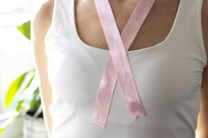 Shot of the woman in the white top against the white wall, with pink ribbon on her neck as a symbol of breast cancer awareness. Concept photo