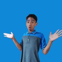 Asian young man with a surprised face facing the camera, with both hands to the side, wearing a blue t-shirt, isolated on a blue background. photo