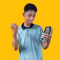 Excited Asian man wearing gray t-shirt pointing to copy space next to holding smart phone, isolated on yellow background. photo